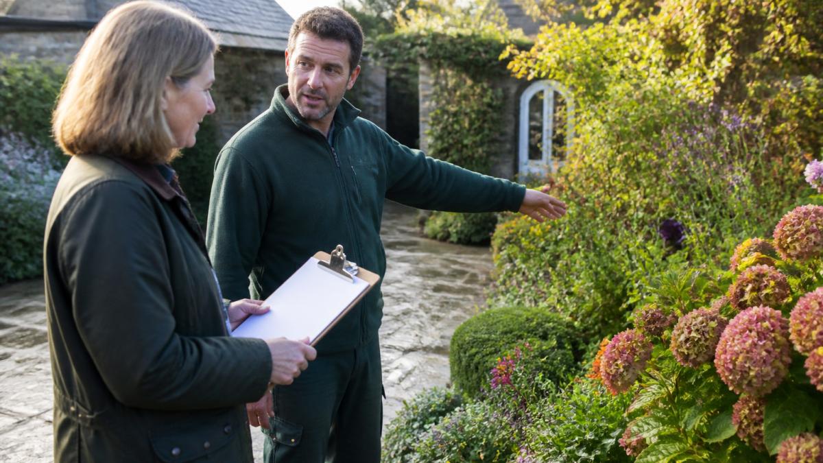A homeowner in a waxed jacket holding a clipboard checklist, talking with a professional gardener who points towards a flower bed, soft morning light across a stone patio